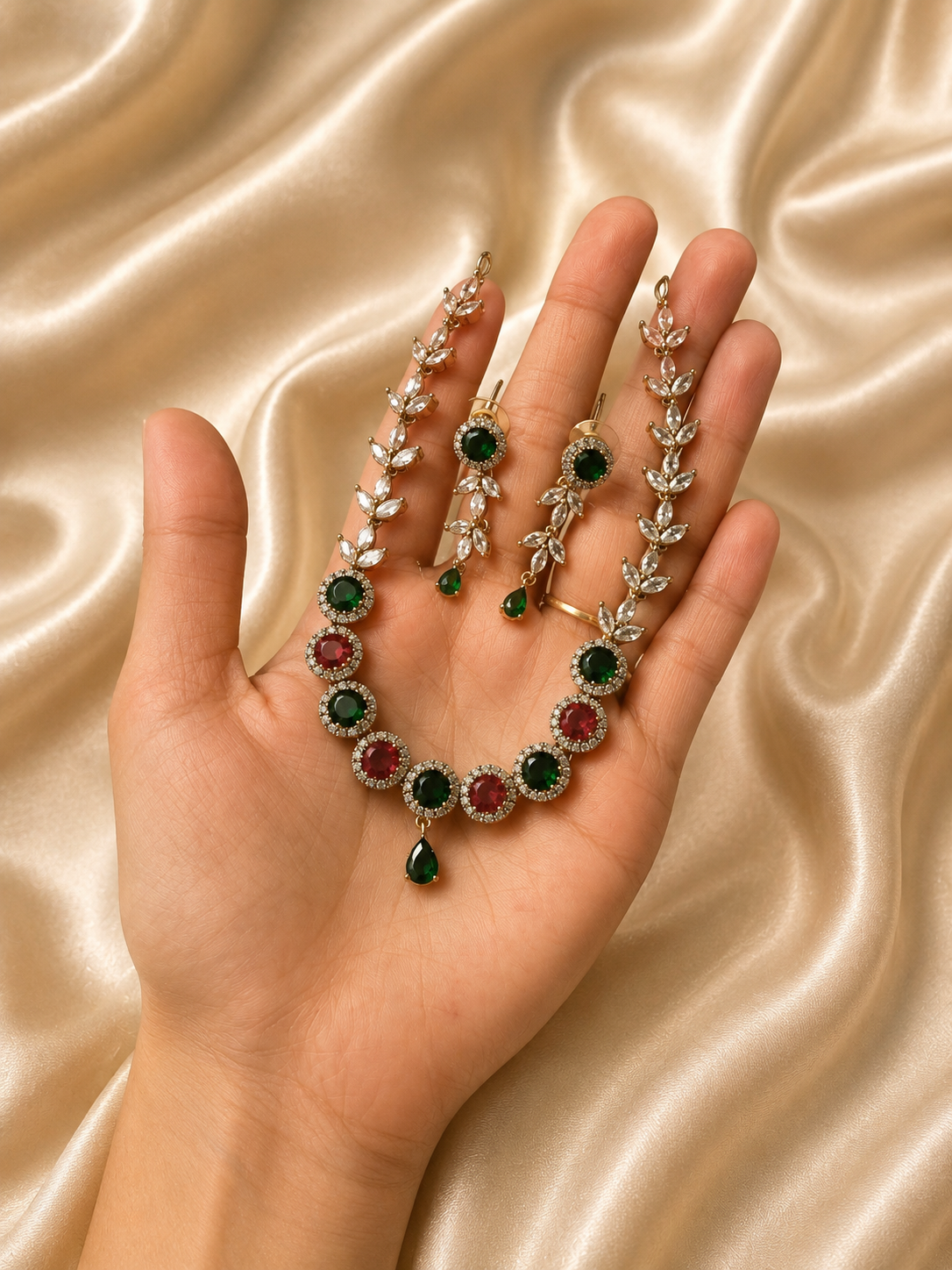 A hand holding a gold-plated necklace set with a chain of clear marquise-cut CZ stones shaped like leaves, leading to alternating round emerald green and ruby red gems, complete with a tiny green teardrop pendant and matching drop earrings against a champagne satin background.