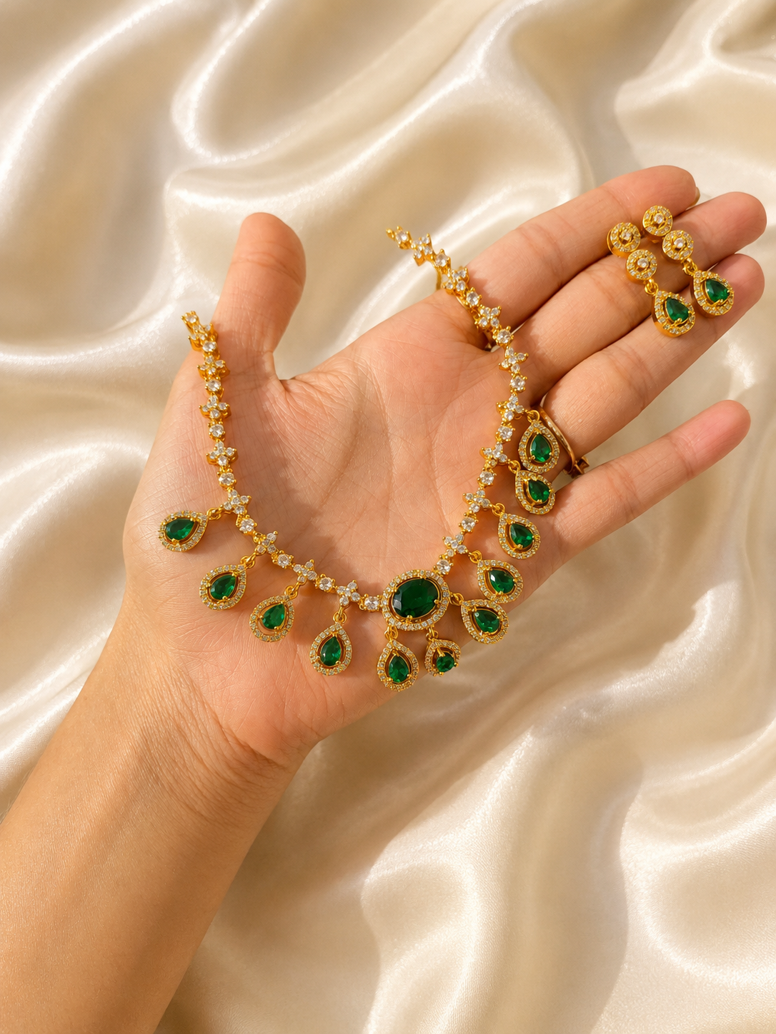 A hand holding a delicate gold-plated choker necklace set featuring dangling teardrop-shaped emerald green stones bordered by white CZ crystals, paired with small matching drop earrings against a cream silk backdrop.