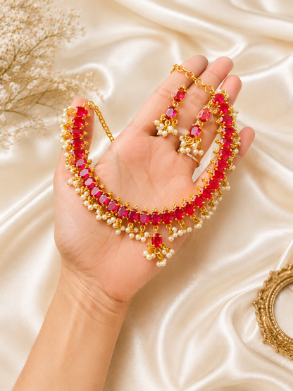 A person holding a gold-plated traditional jewelry set featuring a choker necklace made of a solid row of oval-cut ruby red stones, accented with a fringe of tiny hanging white pearls, complete with matching ruby and pearl drop earrings against a cream satin background.