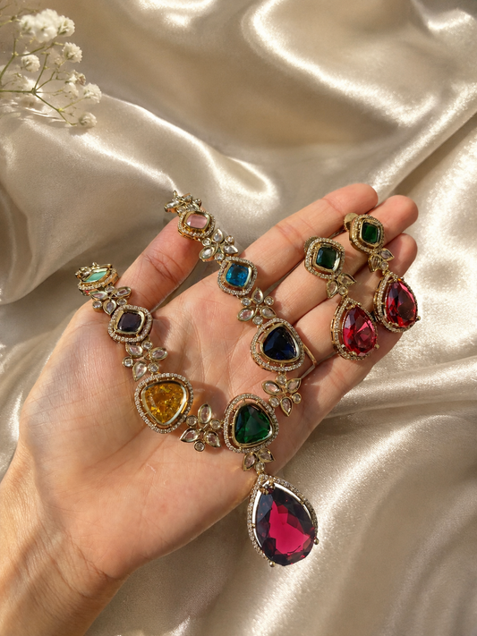 A hand holding an antique Victorian-style jewelry set featuring a collar necklace with large, multi-colored shield and cushion-cut stones in shades of pink, yellow, blue, and green, complete with a prominent teardrop pink pendant and matching drop earrings against a cream silk background.