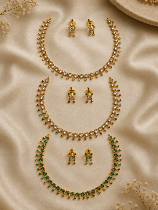 Three minimalist gold-plated choker necklaces arranged vertically on cream satin. The top set features clear crystals with small gold drop spikes; the middle set features alternating clear, red, and green stones; the bottom set features uniform deep green emerald stones. Each set includes matching dangling stud earrings.