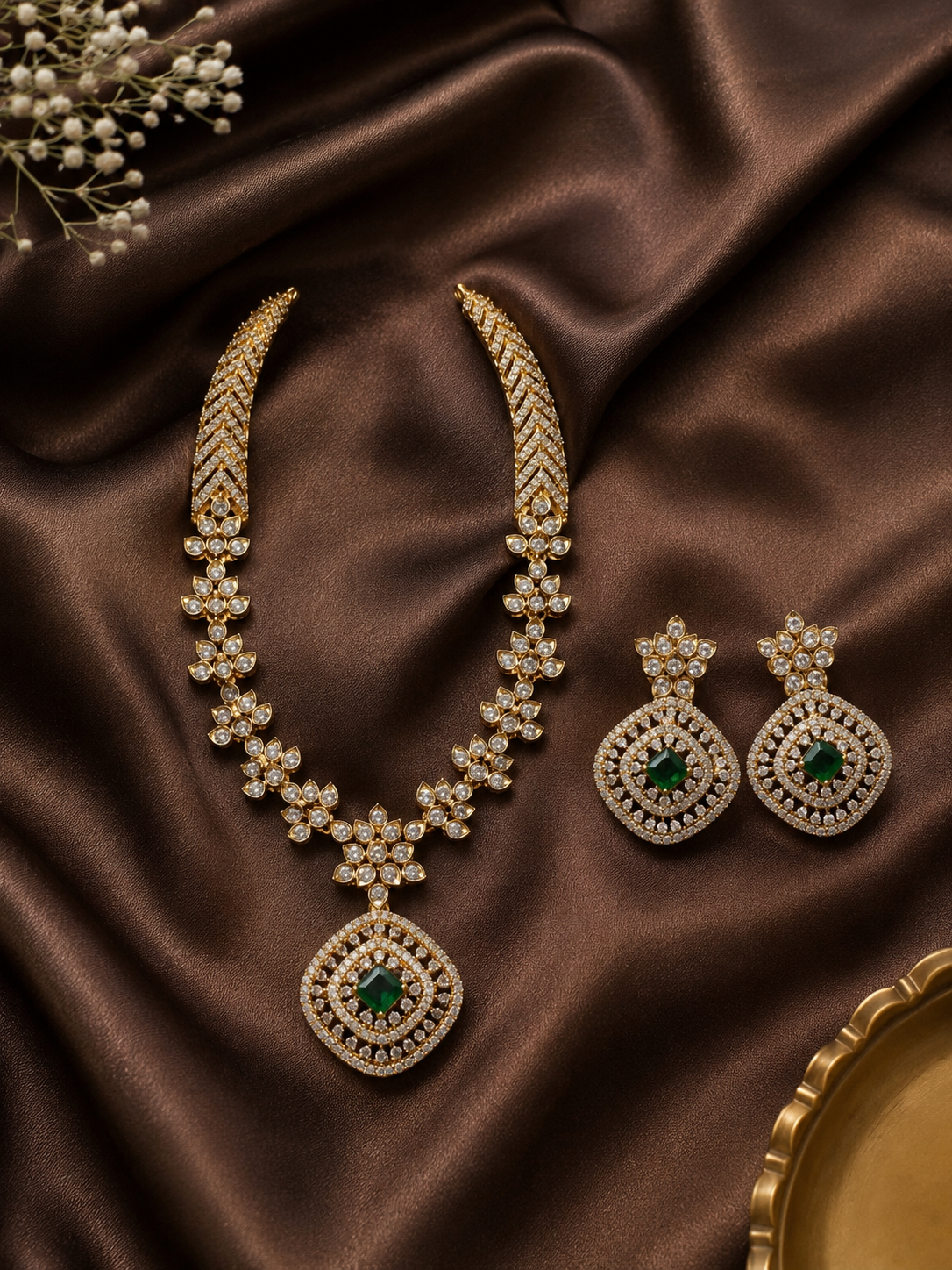 Exquisite gold-plated necklace set featuring a structured, shimmering cubic zirconia collar that transitions into a floral-linked chain and a large diamond-shaped pendant centered with a square green emerald stone. The matching oversized square drop earrings are displayed beside the necklace on a rich, dark brown satin backdrop.
