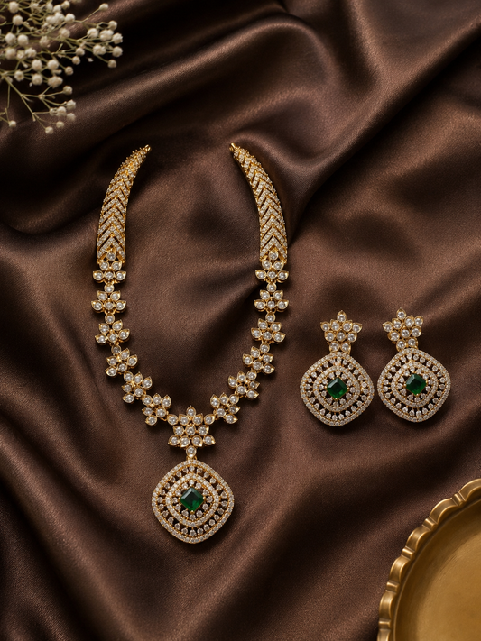 Exquisite gold-plated necklace set featuring a structured, shimmering cubic zirconia collar that transitions into a floral-linked chain and a large diamond-shaped pendant centered with a square green emerald stone. The matching oversized square drop earrings are displayed beside the necklace on a rich, dark brown satin backdrop.