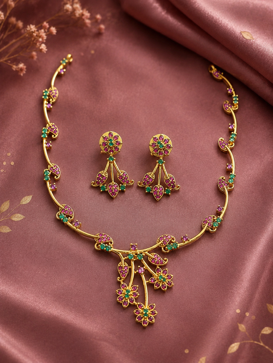 Diya minimalist gold-toned necklace set featuring a delicate asymmetric vine pattern with tiny pink-ruby and emerald-green stones, shown flat on a mauve silk background with matching floral drop earrings.