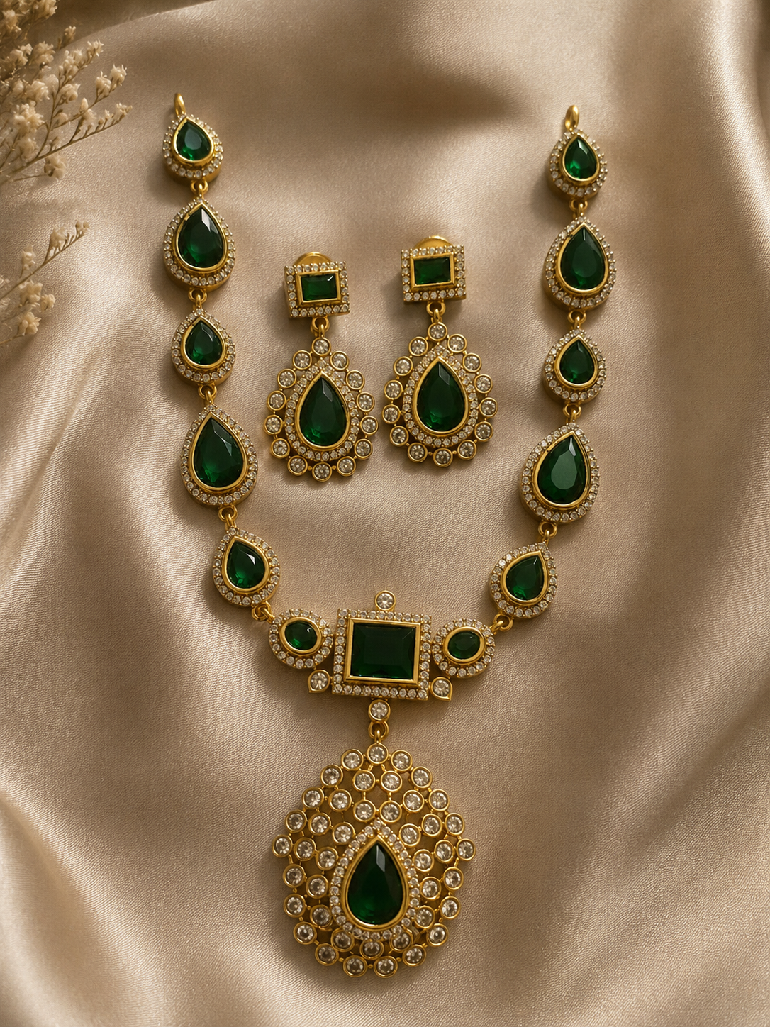 Jyothika Premium jewelry set showcasing a gold-plated necklace with pear-shaped emerald green stones and a large crystal-encrusted teardrop pendant, laid on champagne satin fabric with matching drop earrings.