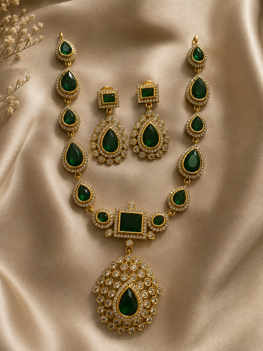 Jyothika Premium jewelry set showcasing a gold-plated necklace with pear-shaped emerald green stones and a large crystal-encrusted teardrop pendant, laid on champagne satin fabric with matching drop earrings.