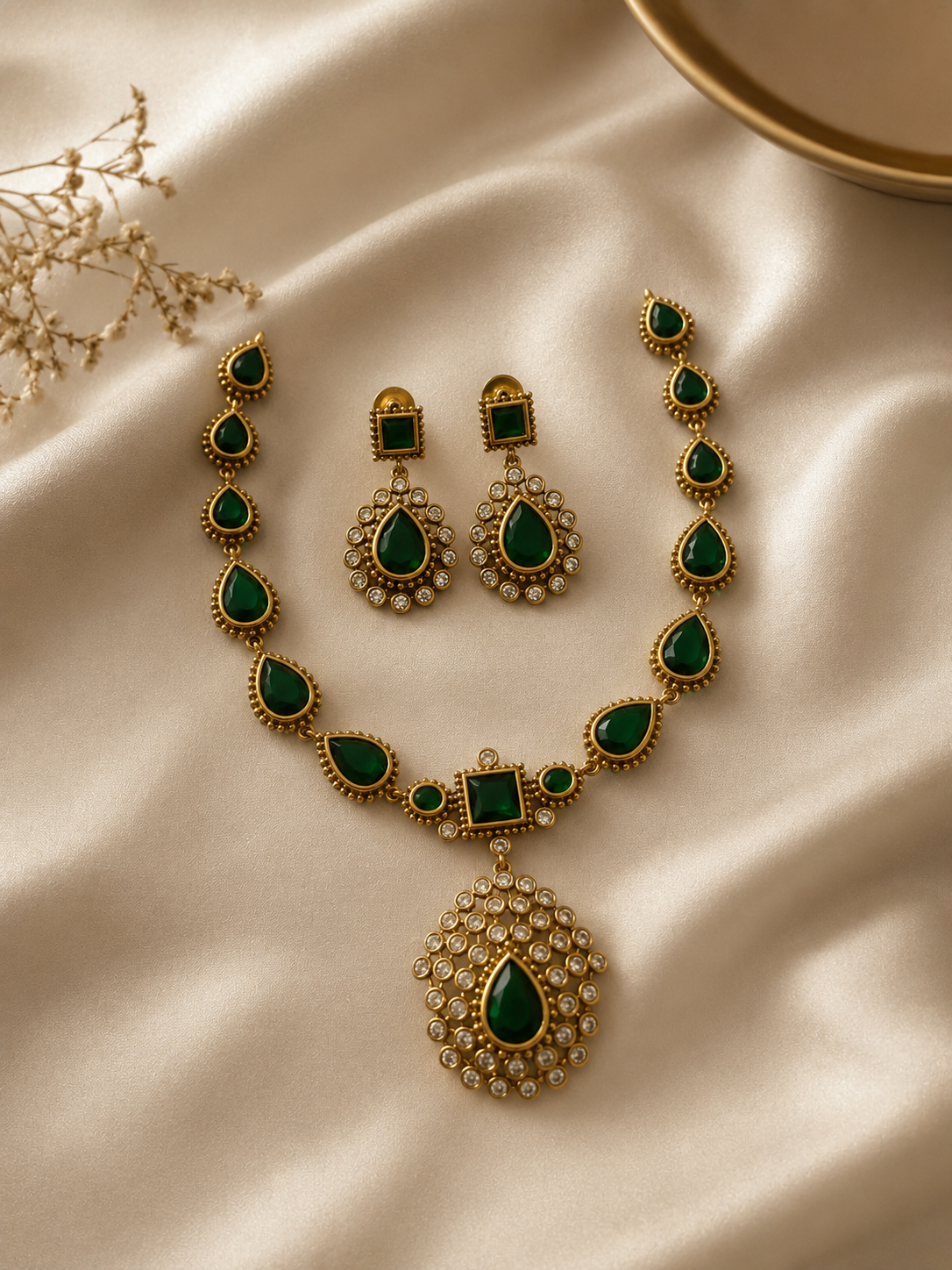 Antique-finish jewelry set showcasing a matte gold-plated necklace with pear-shaped emerald green stones and a large crystal-encrusted teardrop pendant, displayed on cream satin fabric with matching square-stud drop earrings.