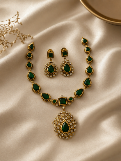 Antique-finish jewelry set showcasing a matte gold-plated necklace with pear-shaped emerald green stones and a large crystal-encrusted teardrop pendant, displayed on cream satin fabric with matching square-stud drop earrings.