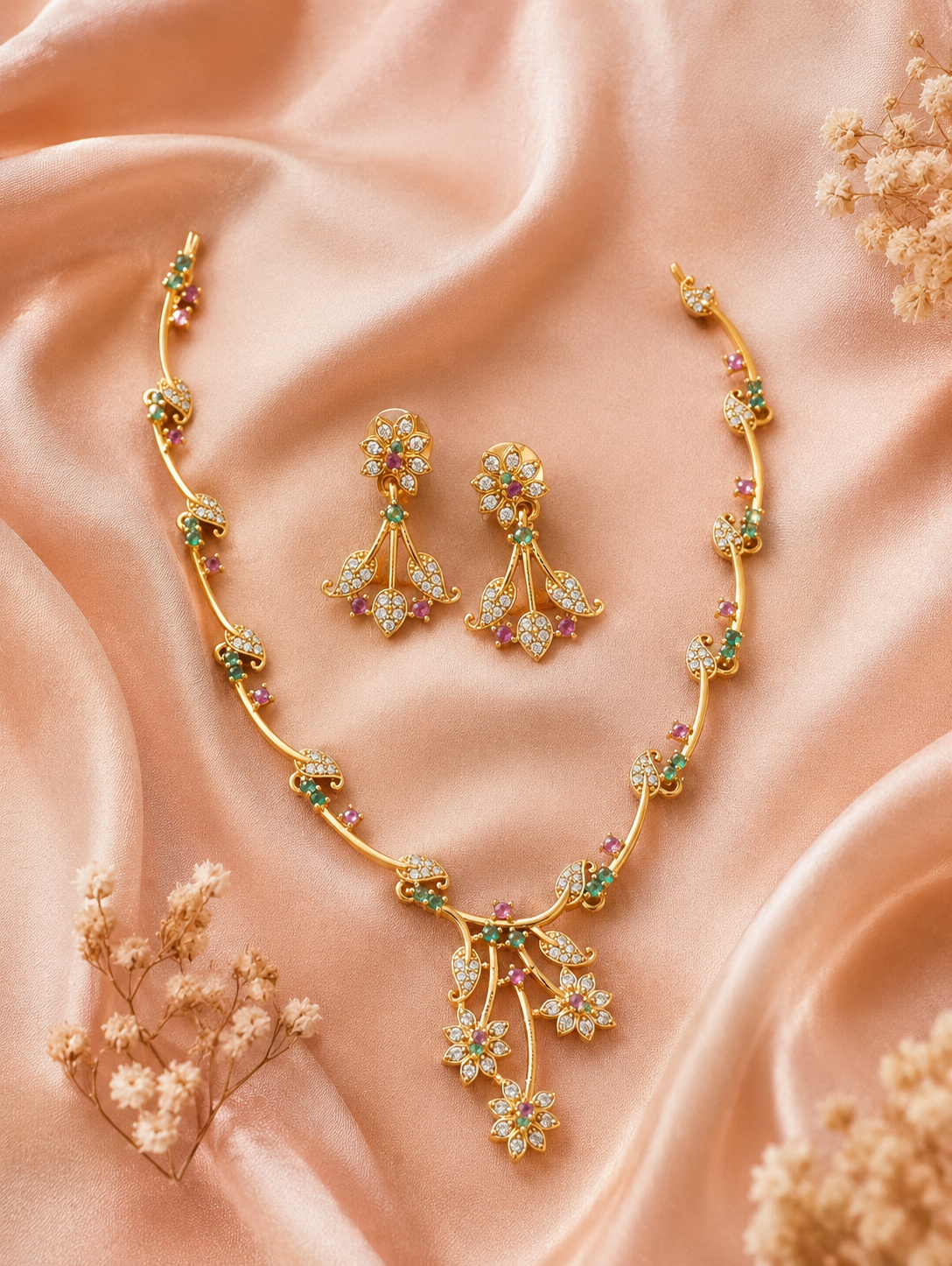 Miraya minimalist gold-toned necklace set featuring a delicate asymmetric vine pattern with tiny clear crystals, ruby-pink, and emerald-green accent stones, laid flat on soft peach satin fabric with matching floral drop earrings.