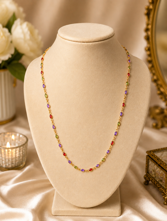 Anagha minimalist gold-plated mala necklace chain featuring a continuous row of small bezel-set oval crystals in red, purple, light green, and dark green, displayed elegantly on a cream jewelry bust stand.
