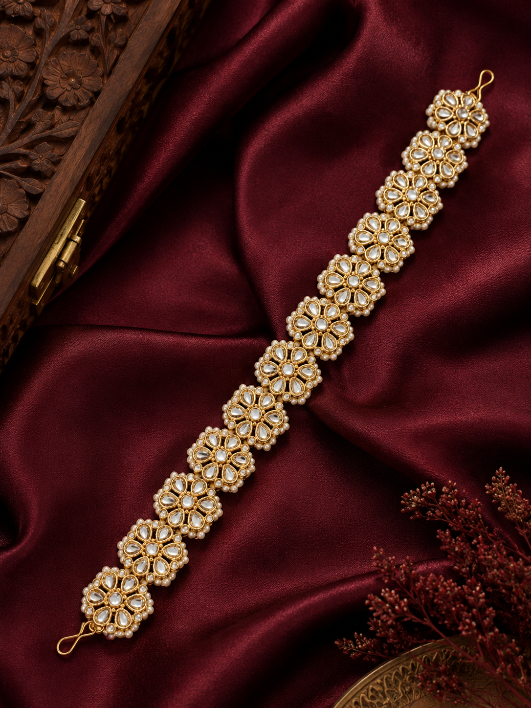 Traditional Indian gold-plated sheeshphool hair band featuring a continuous row of circular floral motifs set with glistening Kundan stones and tiny white pearls, shown laid flat on rich maroon satin fabric and modeled on a woman's hair.