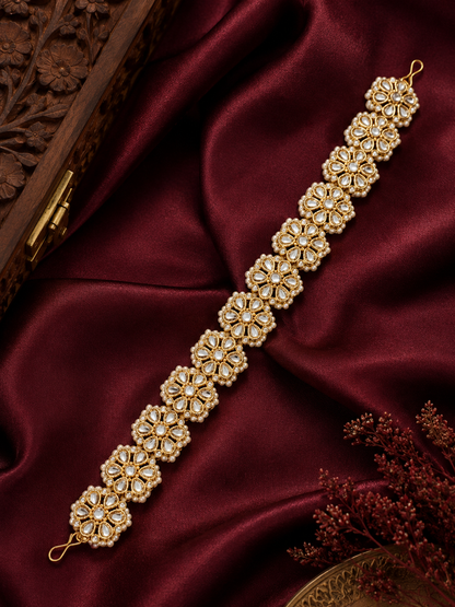 Traditional Indian gold-plated sheeshphool hair band featuring a continuous row of circular floral motifs set with glistening Kundan stones and tiny white pearls, shown laid flat on rich maroon satin fabric and modeled on a woman's hair.