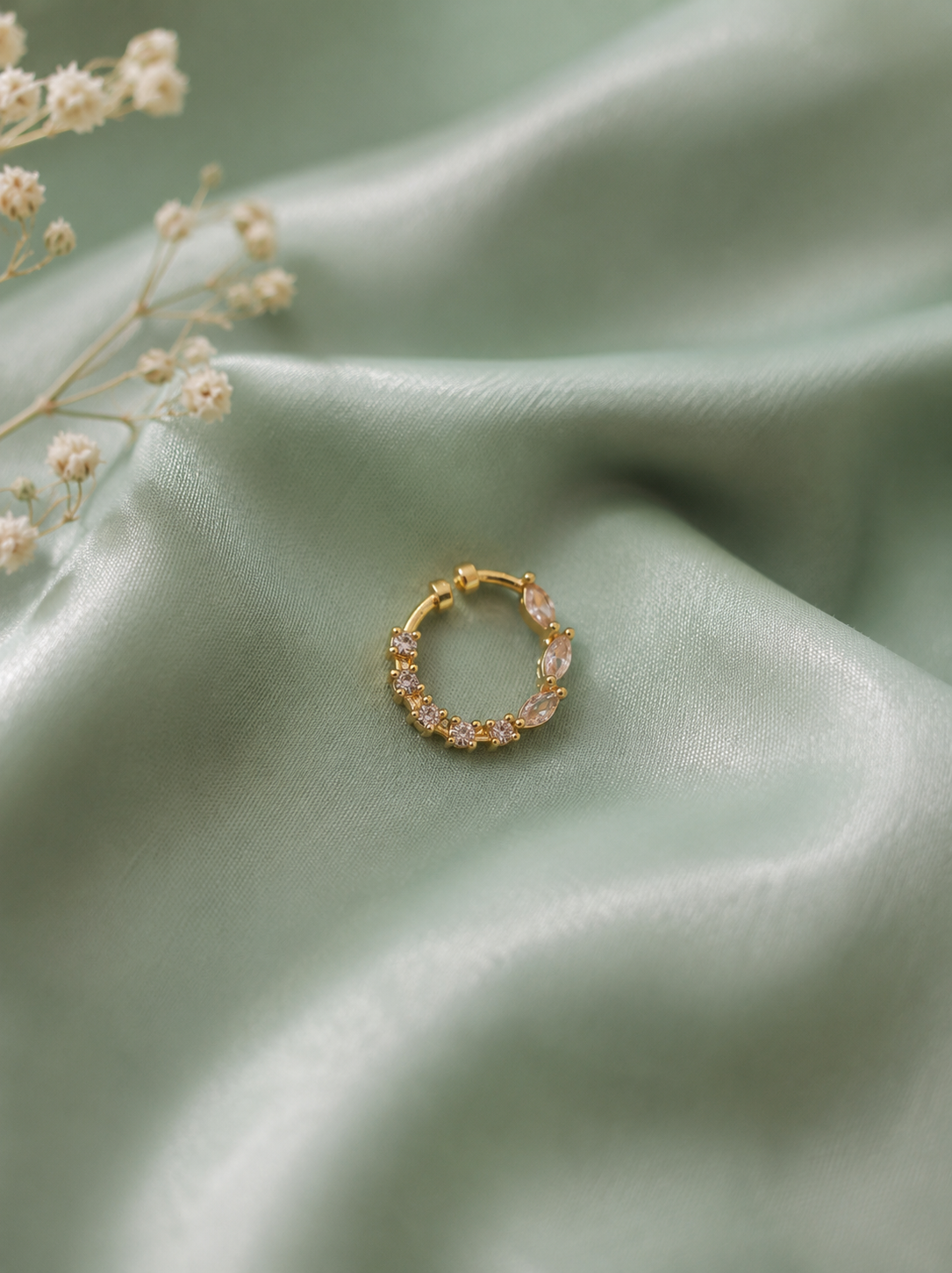 A delicate gold-tone festive nose ring adorned with clear white stones, resting on a soft, rippled sage green satin fabric next to small white dried baby's breath flowers.