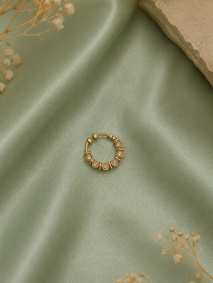 A gold-tone diamond septum ring featuring a crescent of round bezel-set clear crystals, placed on a draped sage green satin fabric with delicate dried baby's breath flowers.
