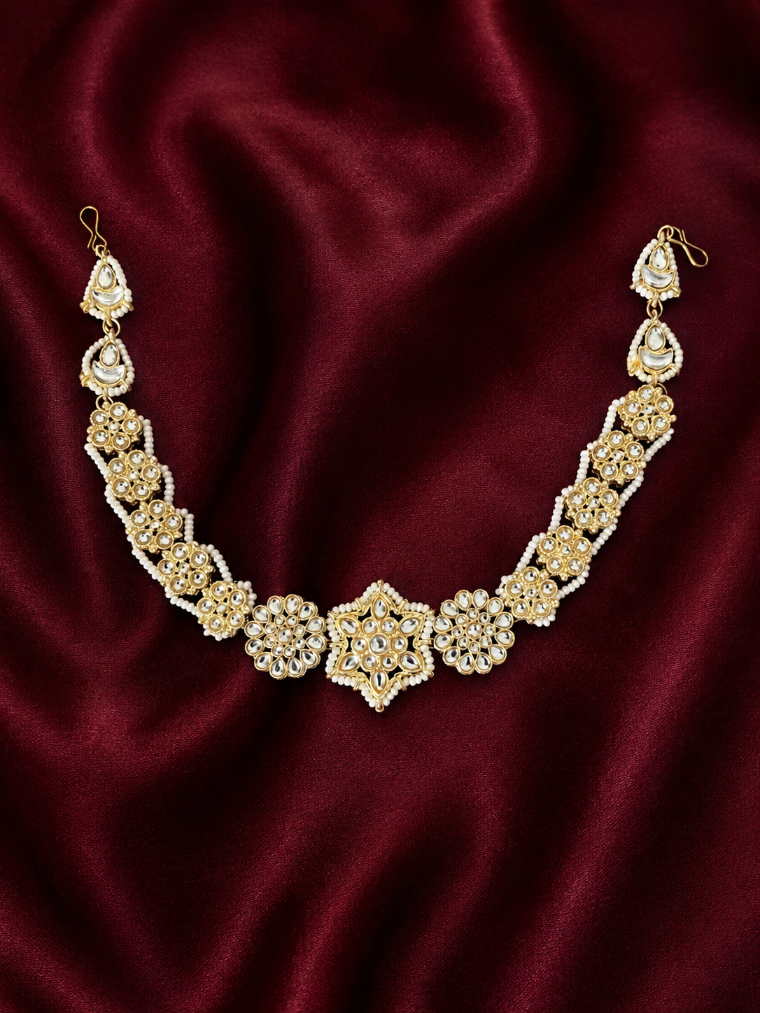 Heirloom-style gold-plated Indian sheeshphool headpiece featuring a central star-shaped Kundan medallion flanked by pearl-edged floral patterns, shown on a rich dark red satin backdrop and worn by a smiling model.