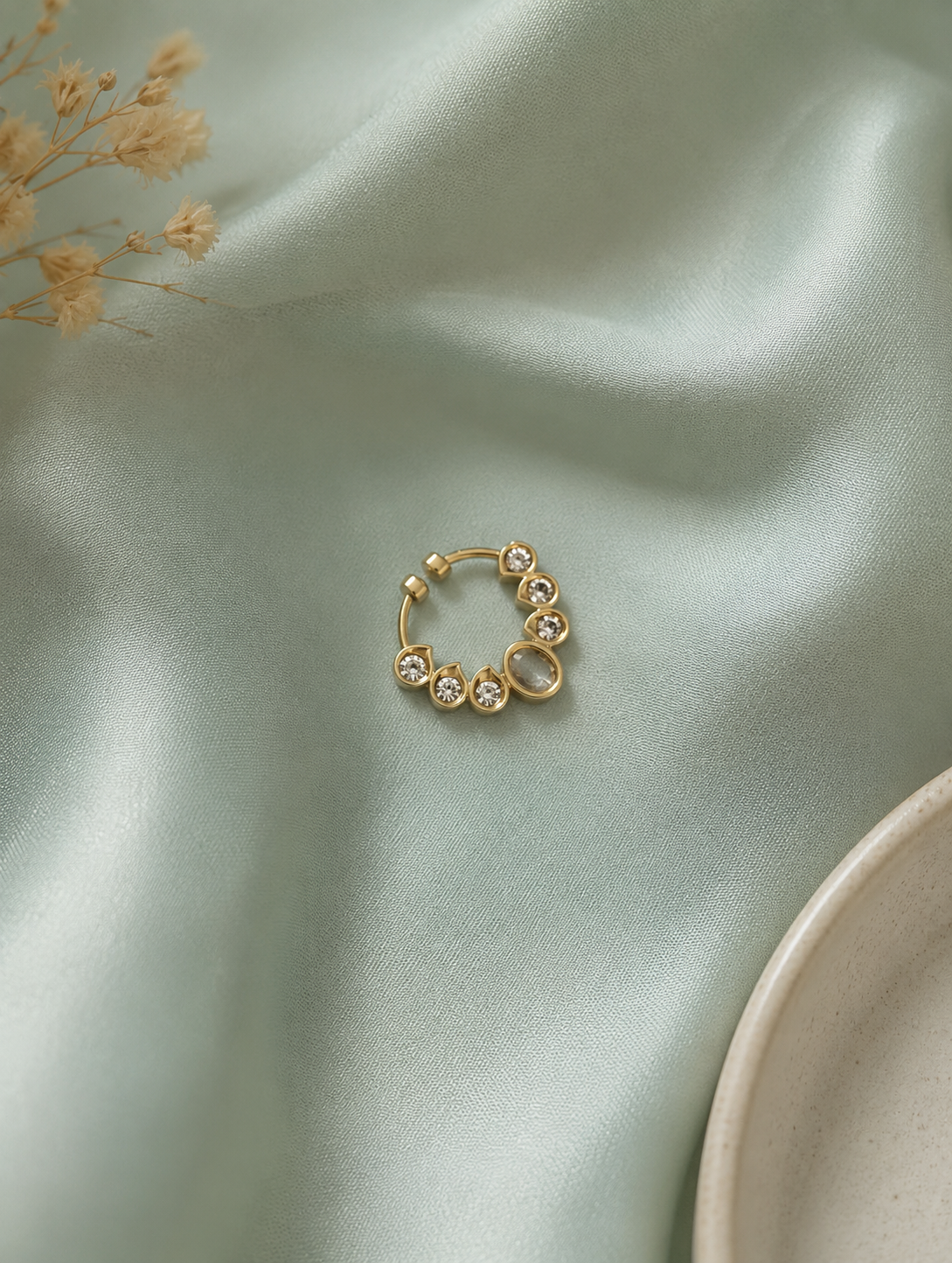 A decorative gold-tone septum ring designed with a prominent bezel-set oval stone surrounded by sparkling round crystals, resting on elegant sage green satin fabric near dried floral details.