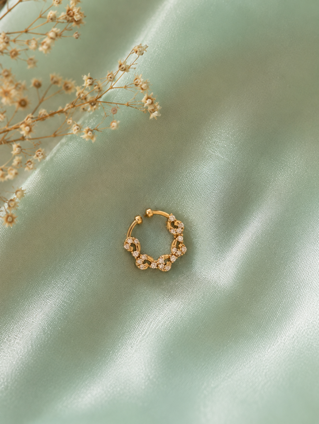 An intricate gold-tone septum ring featuring a scalloped, wavy floral filigree design encrusted with tiny white crystals, centered on a draped sage green satin fabric with sprigs of dried baby's breath.