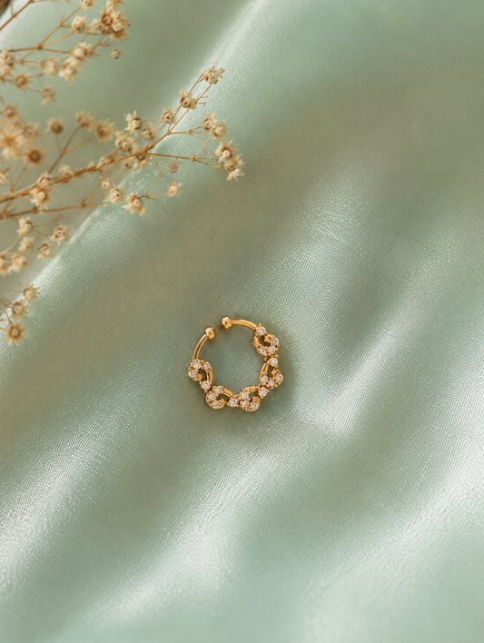 An intricate gold-tone septum ring featuring a scalloped, wavy floral filigree design encrusted with tiny white crystals, centered on a draped sage green satin fabric with sprigs of dried baby's breath.