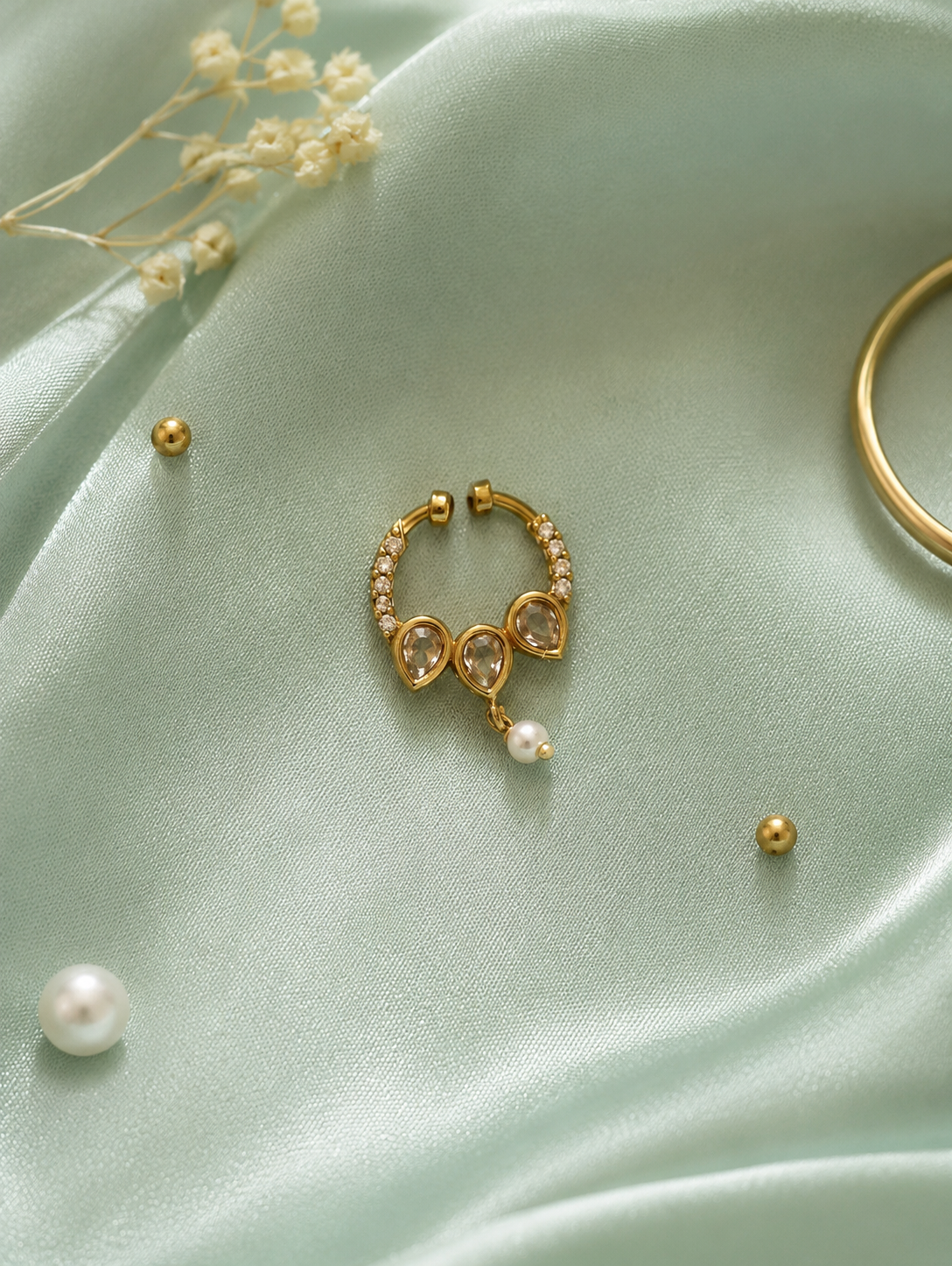 An elegant gold-tone diamond septum ring featuring three teardrop-shaped clear crystals and a small dangling pearl, displayed on a soft, light-green silk fabric background.