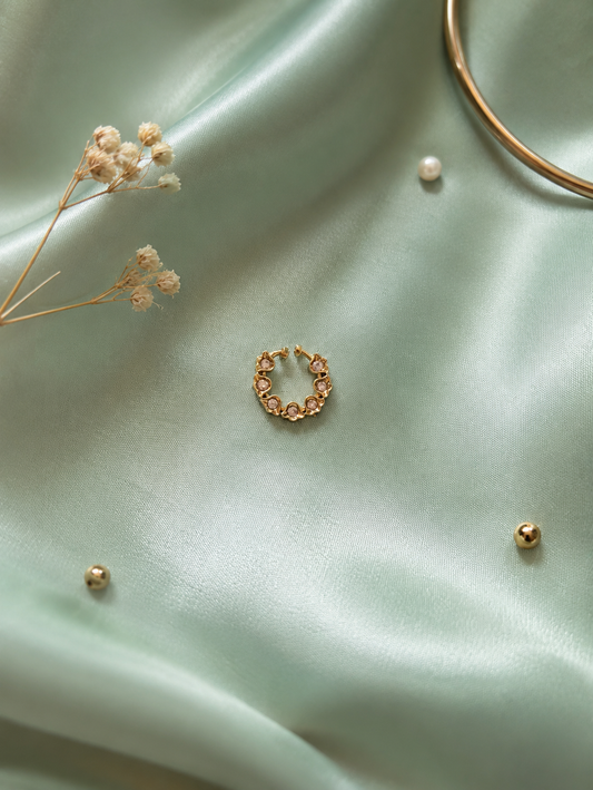 A dainty floral gold-tone septum hoop ring featuring a crown of small flower patterns inlaid with tiny clear crystals, displayed centrally on a shimmering light-green satin fabric background.