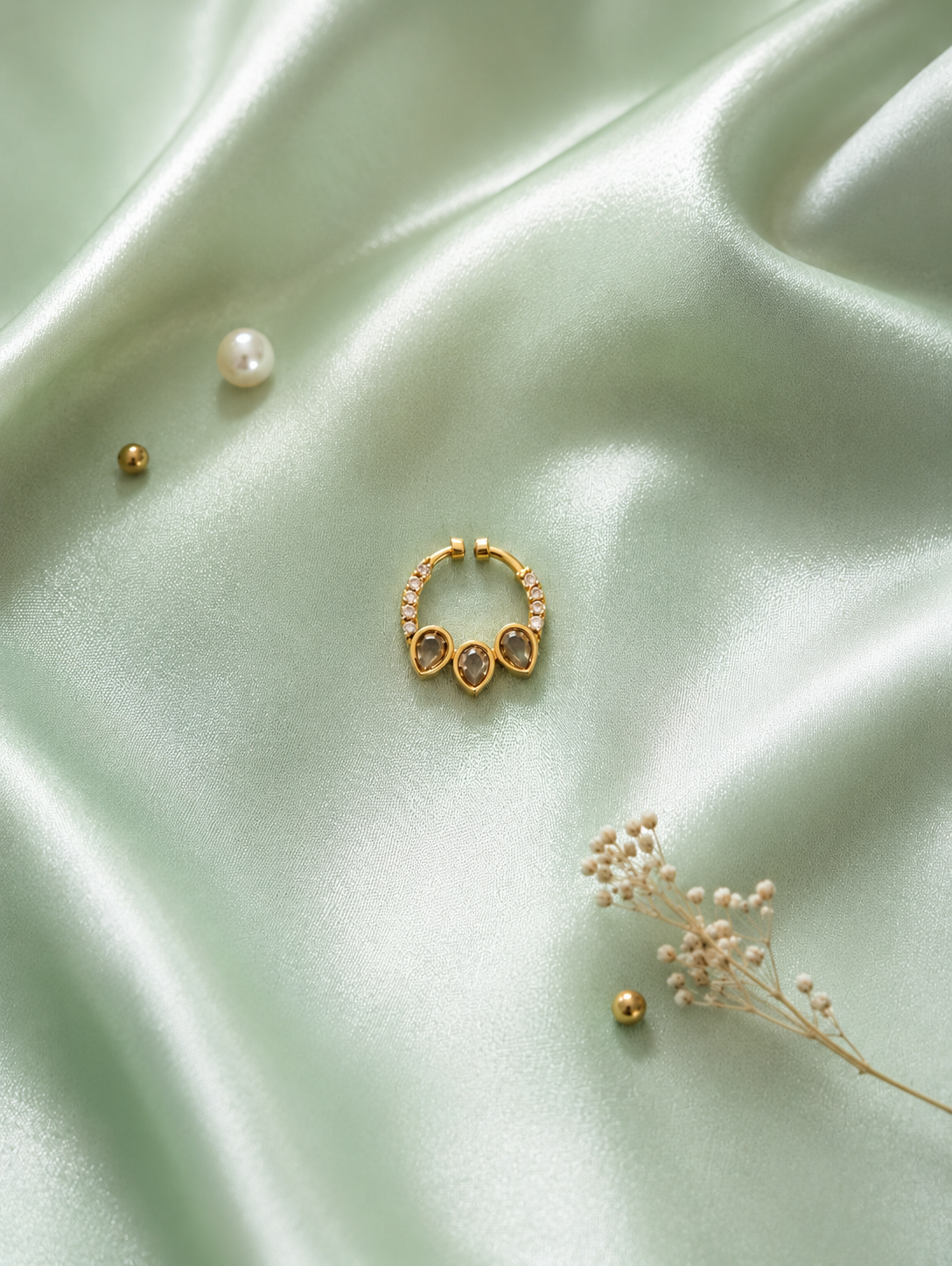 A gold-tone diamond septum clicker ring featuring a curved band lined with small round clear crystals and three central hanging teardrop-shaped smoky grey stones, displayed flat on shimmering sage green satin fabric with a pearl accent.