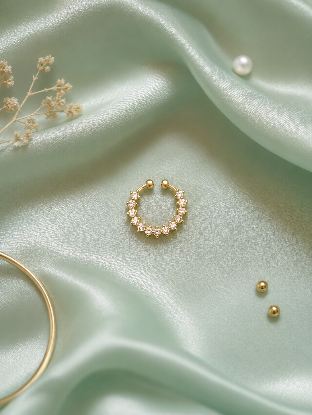 A gold-tone crystal septum ring and ear cuff featuring a non-piercing crescent band lined with clustered round clear crystals in prong settings, displayed flat on shimmering sage green satin fabric with a gold bangle and pearl accent.