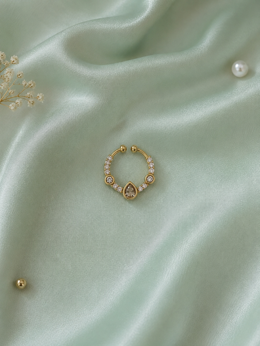 Elegant gold-toned septum ring featuring a central teardrop crystal and surrounding white diamond accents, displayed on a textured sage green silk background.