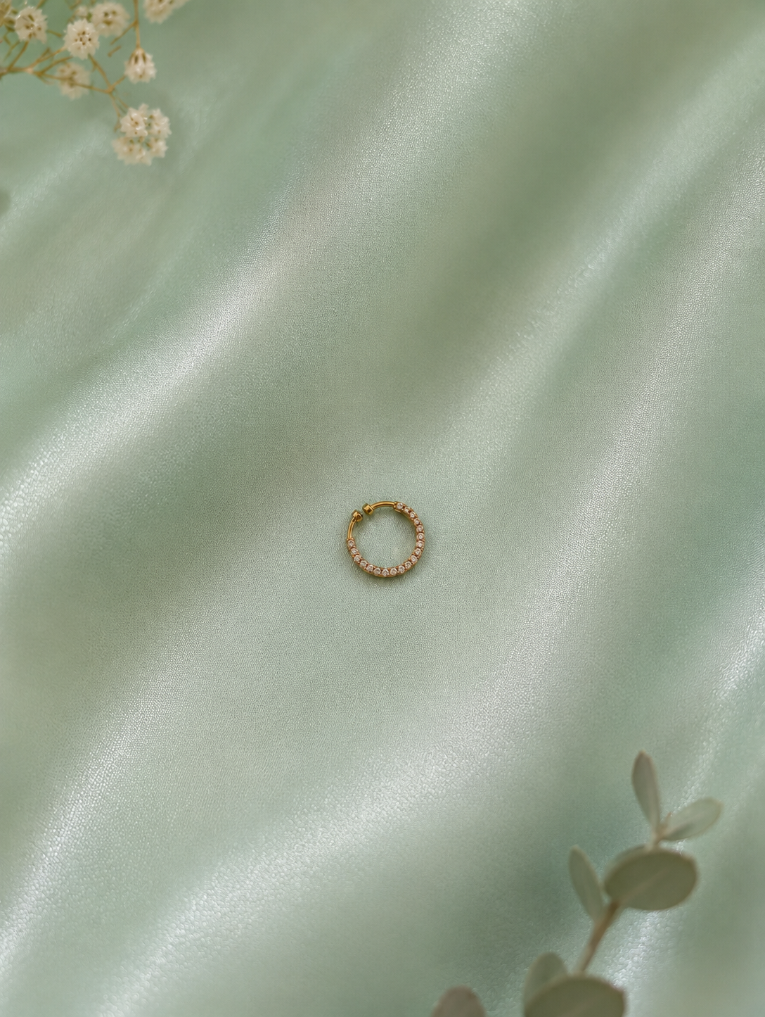 Minimalist overhead shot of a circular gold-toned pave crystal septum ring centered on a shimmering mint green satin fabric background, accented by subtle floral elements.