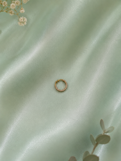 Minimalist overhead shot of a circular gold-toned pave crystal septum ring centered on a shimmering mint green satin fabric background, accented by subtle floral elements.