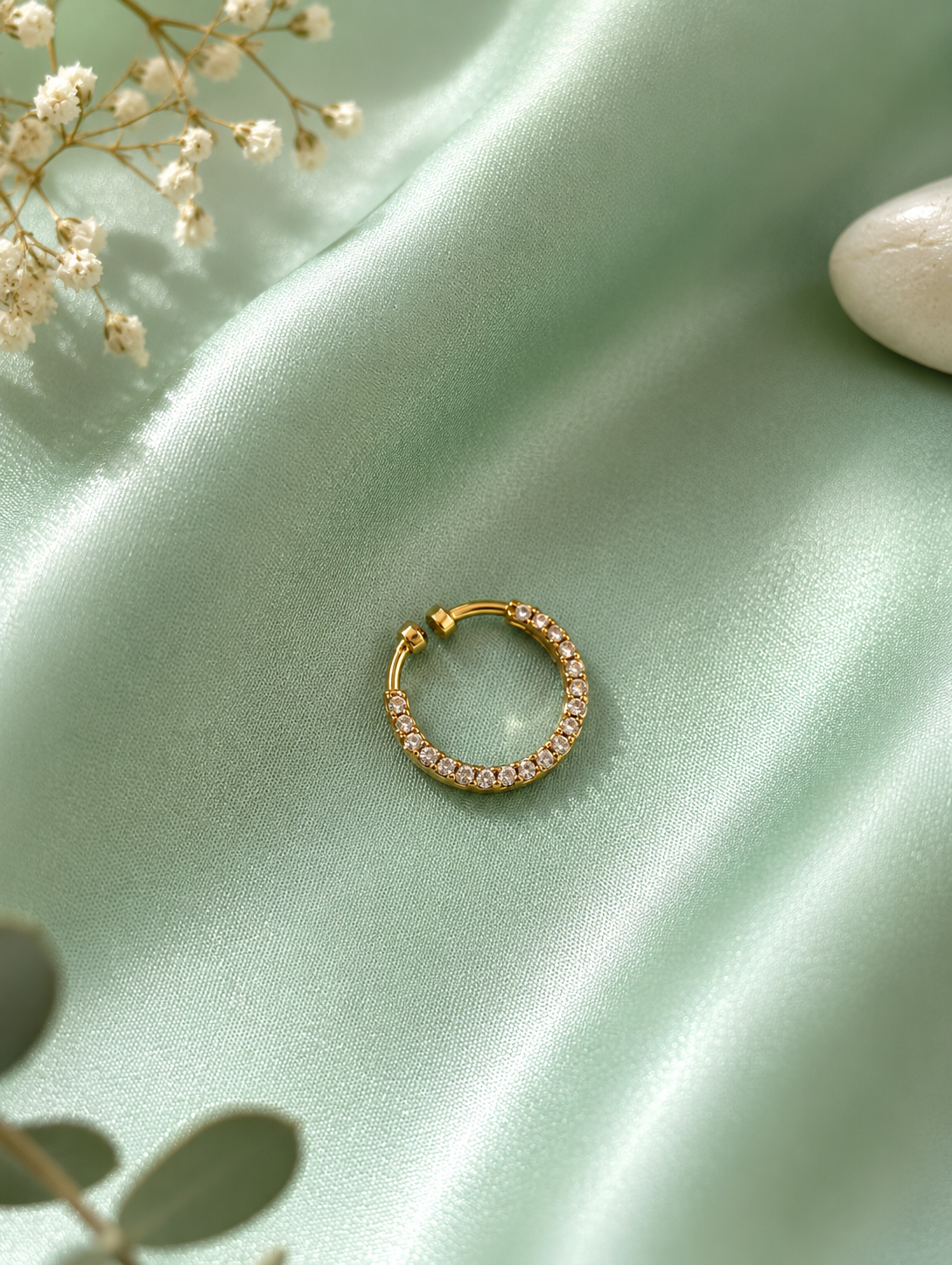 A close-up view of a gold-toned septum ring lined with small, sparkling pave crystals, displayed on a draped sage green silk cloth next to a white stone and delicate white baby's breath flowers.