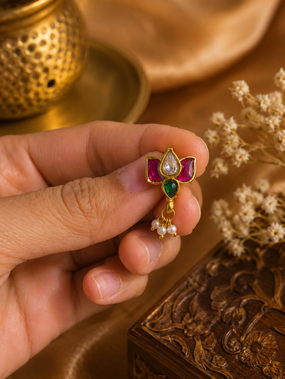 A person holding the SP013 traditional Indian Jadau Kundan lotus nose pin between their fingers, showcasing the intricate gold setting, vibrant pink, clear, and green gemstones.
