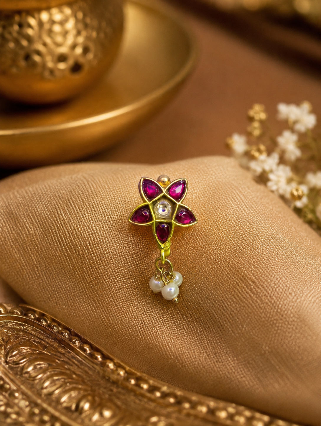 "An exquisite gold star-shaped nose pin with red stones and dangling white pearls resting on a soft, beige satin fabric."