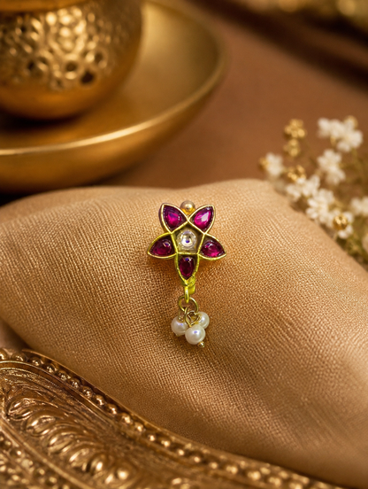 "An exquisite gold star-shaped nose pin with red stones and dangling white pearls resting on a soft, beige satin fabric."