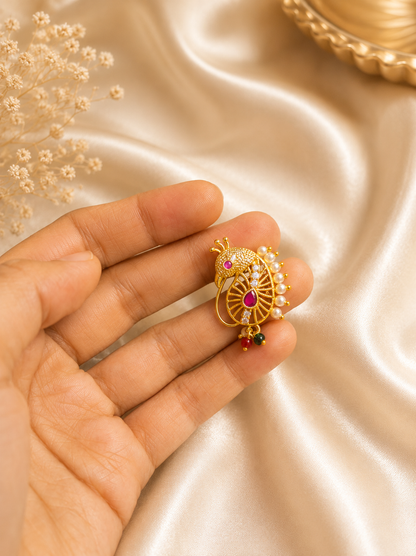 A hand holding the traditional gold Maharashtrian Peshwai peacock Nath against a soft, elegant champagne silk fabric background with subtle decorative elements, showcasing its fine details and pearl embellishments.