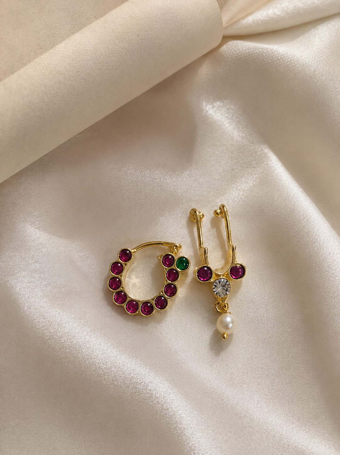 Gold-plated traditional Indian nath nose rings with ruby pink stones, green accents, and a hanging white pearl drop on a cream satin background.