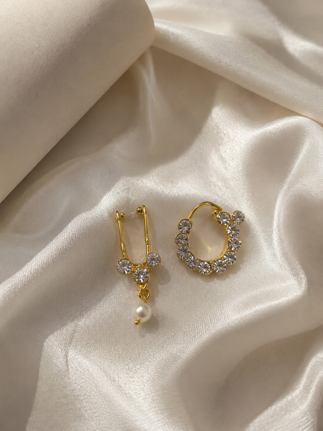 A gold-plated traditional Indian nath and septum combo jewelry set studded with circular clear white crystals and a hanging pearl drop, placed on a white satin fabric backdrop.