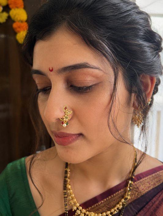A close-up portrait of a woman wearing traditional Indian jewelry, featuring a gold nose ring (Hastapundra) with a small red and green stone accent, a gold necklace, and earrings, paired with a green and maroon sari.