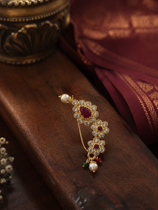 A gold-toned traditional Indian bindi pin featuring a central red ruby stone, surrounded by white stones, with small pearl and red-green bead accents, displayed on a dark wooden surface.