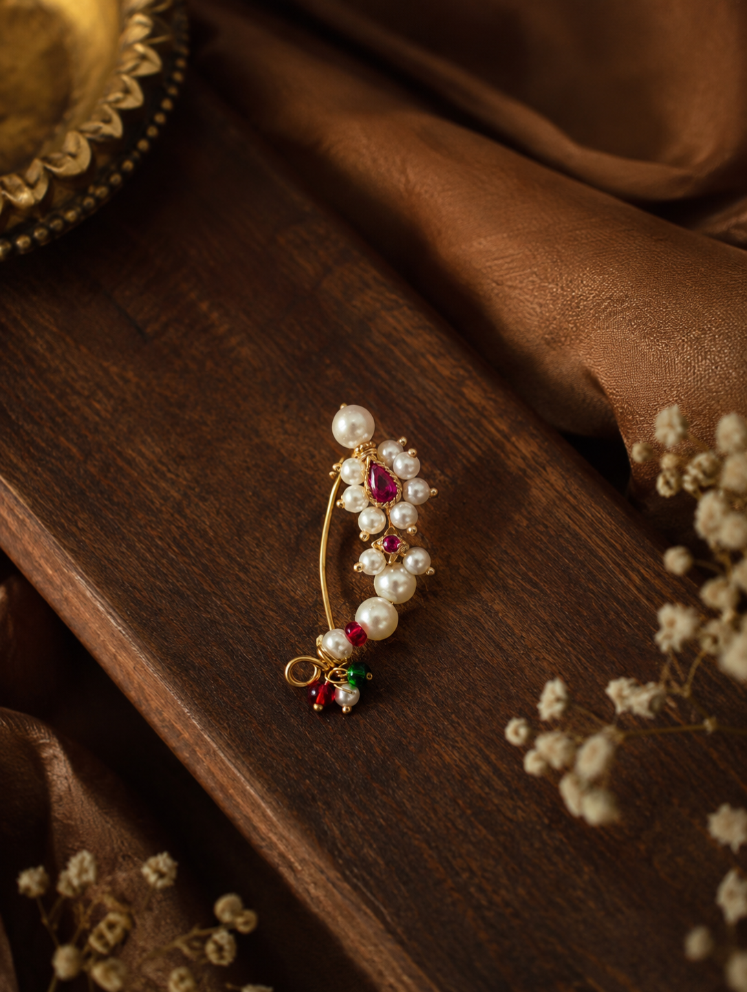 A gold-toned traditional Indian style hair pin adorned with white pearls, pink and green gemstones, and a red teardrop stone, displayed on a dark wooden surface with brown fabric and dried flowers.


