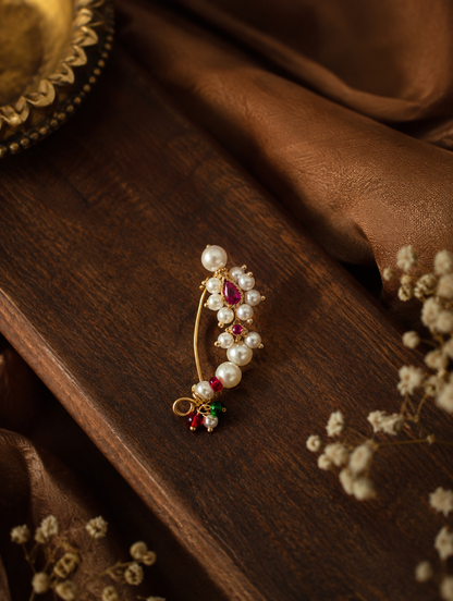 A gold-toned traditional Indian style hair pin adorned with white pearls, pink and green gemstones, and a red teardrop stone, displayed on a dark wooden surface with brown fabric and dried flowers.

