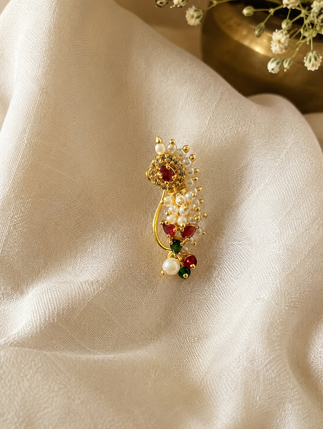 A gold-toned statement brooch featuring white faux pearls, red gemstone accents, and small green stones, displayed on draped cream-colored fabric.