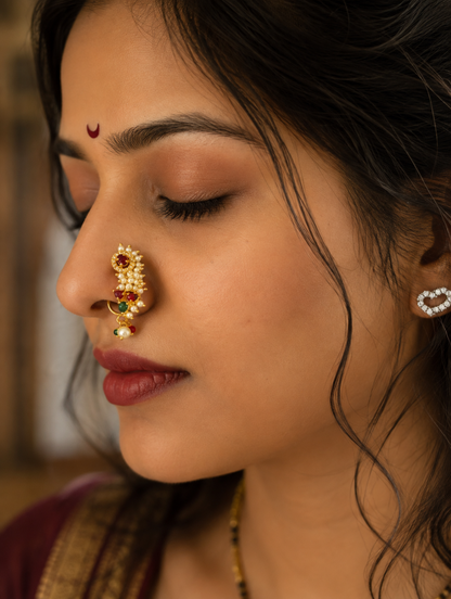 A close-up side profile of a woman wearing a gold nose ring (nath) adorned with red and green stones, along with a matching heart-shaped stud earring.