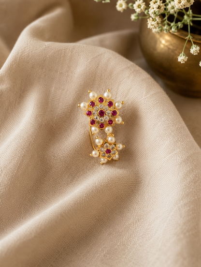 A gold-plated ring featuring two floral clusters of white pearls and red gemstones, displayed on draped beige fabric with a decorative gold pot in the background.