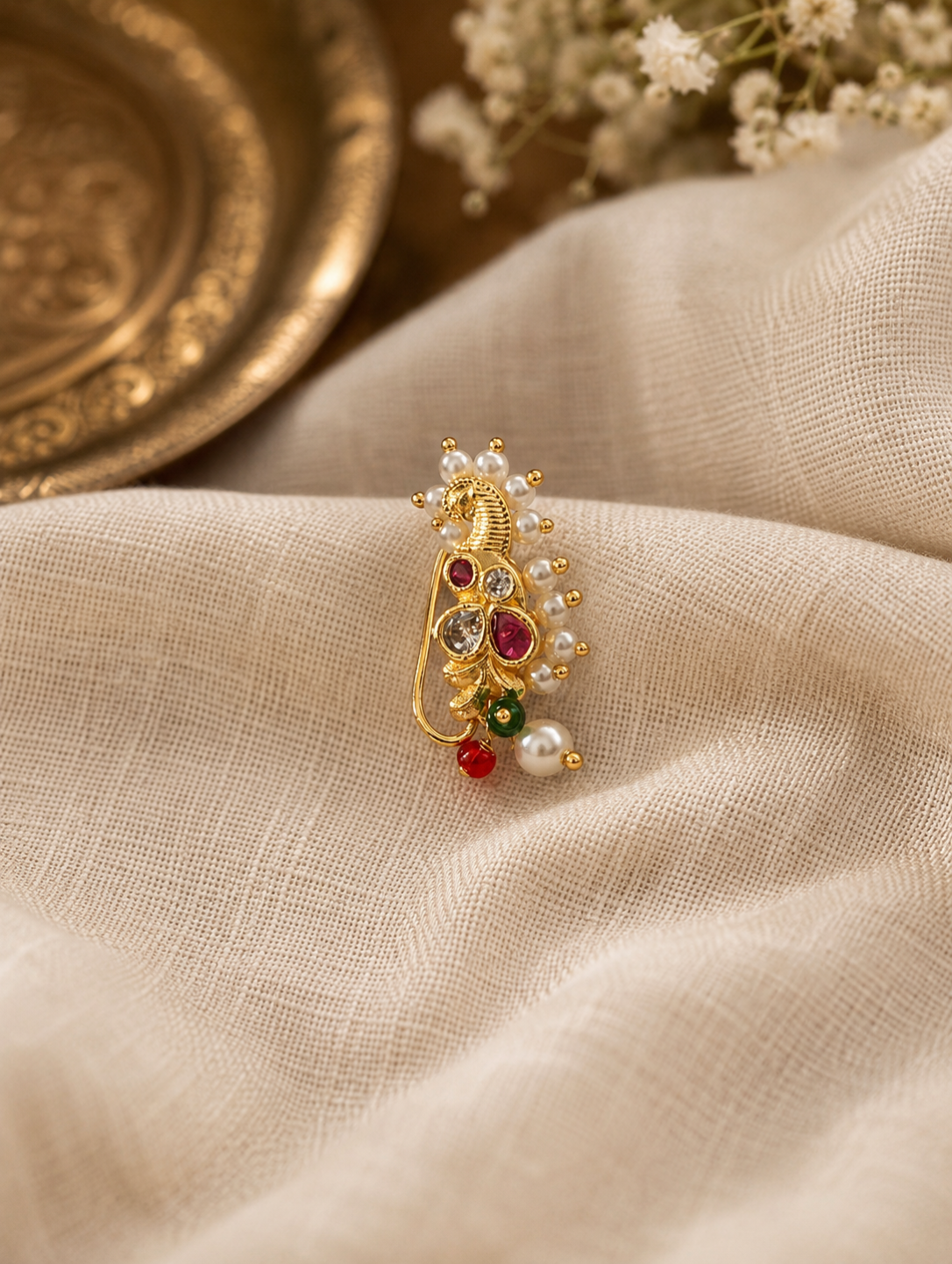 A gold-toned traditional Indian jewelry piece, likely a brooch or decorative charm, featuring a peacock motif with red and green stones and white pearl accents, resting on a textured beige fabric.