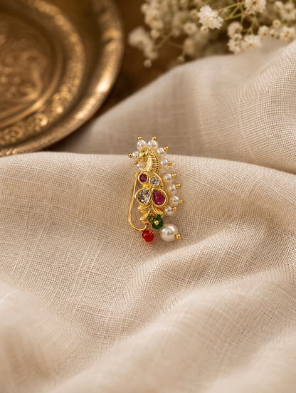 A gold-toned traditional Indian jewelry piece, likely a brooch or decorative charm, featuring a peacock motif with red and green stones and white pearl accents, resting on a textured beige fabric.