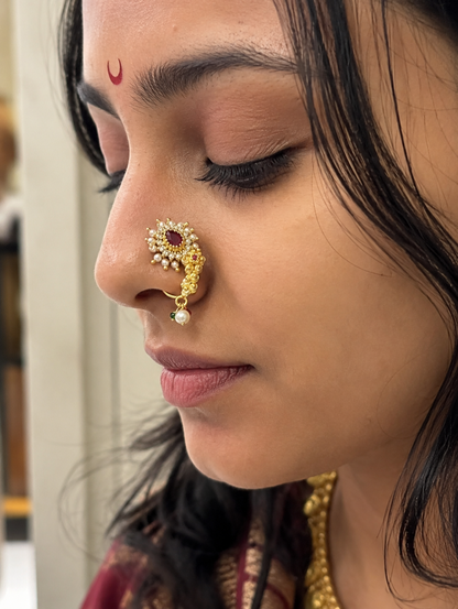 A close-up side profile of a woman wearing a gold crescent-shaped nose ornament (ushnōgliya) adorned with small white stones and a central red gemstone, with a red bindi on her forehead.