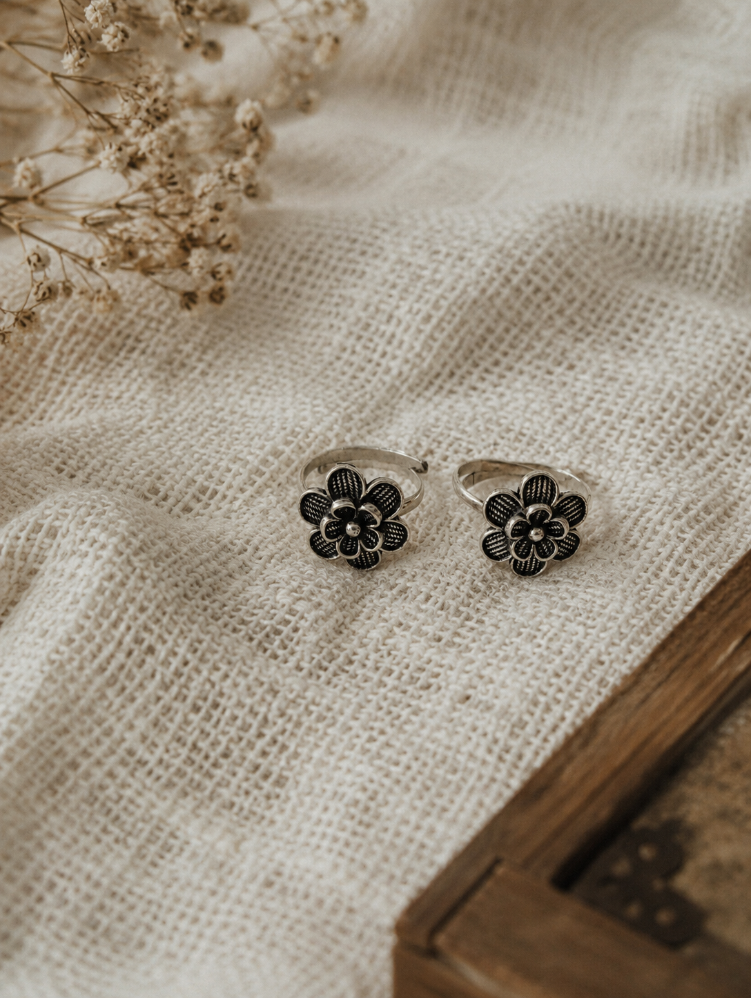 Two silver floral rings on a textured beige fabric surface with dried flowers.