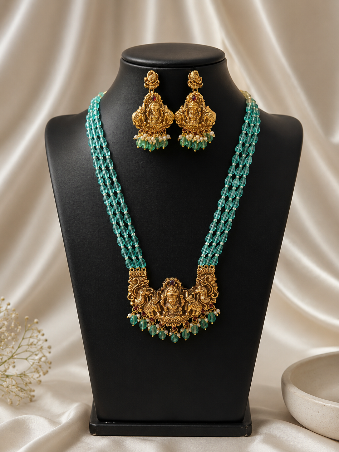 Gold necklace and earrings with green beads on a black mannequin against a white fabric background