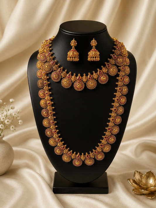 A complete South Indian bridal temple jewelry set including a short choker, a matching long haram necklace with ruby-studded circular floral motifs, and coordinating jhumka earrings on a black display stand.