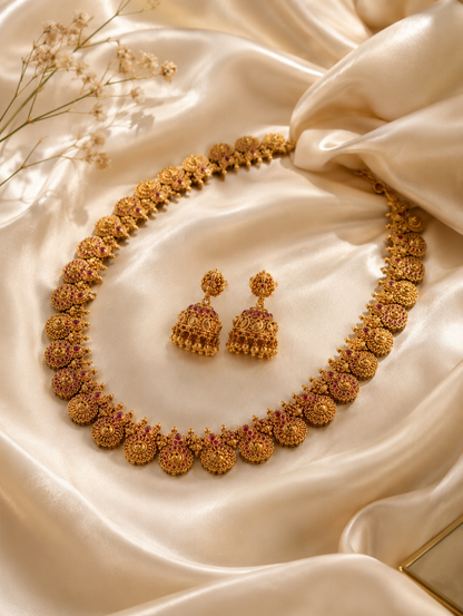 An antique gold-toned temple jewelry set featuring a long haram necklace made of continuous circular floral motifs accented with rubies, paired with matching hanging jhumkas on a black display bust.
